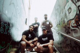 4 dunkel gekleidete Männer auf einer Treppe, an den Seiten sind Wände mit Grafitti, im Hintergrund ein überbelichteter weißer Himmel