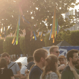 Festivalbesucher:innen im SOnnenuntergang vor einem Baum mit bunter Dekoration