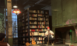 Ein Mann mit roter Kopfbedeckung sitzt auf einer B&uuml;hne vor einem B&uuml;cherregal