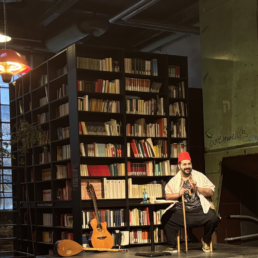 Ein Mann mit roter Kopfbedeckung sitzt auf einer Bühne vor einem Bücherregal