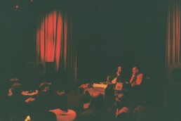 Zwei Herren sitzen auf einer rot-beleuchteten Bühne von Publikum umringt.