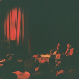 Zwei Herren sitzen auf einer rot-beleuchteten Bühne von Publikum umringt.