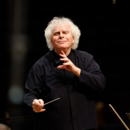 Sir Simon Rattle dirigiert lächelnd mit geschlossenen Augen und hält den Taktstock in der rechten Hand.