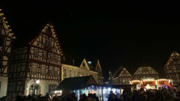 Gemütlich leuchtende Fachwerkhäuser auf einem Marktplatz, im Vodergrund Buden eines Weinachtsmarktes.