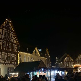 Gemütlich leuchtende Fachwerkhäuser auf einem Marktplatz, im Vodergrund Buden eines Weinachtsmarktes.