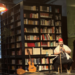 Muawia Harb sitzt auf einer kleinen Bühne mit Instrumenten, hinter ihm steht ein großes Bücherregal
