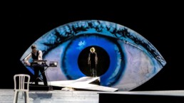 Eine Bühne mit zwei Personen, im Hintergrund ein riesiges blaues Auge