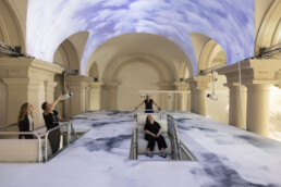 Ein Saal mit großen Säulen auf dessen Decke und Boden Wolken projiziert sind.