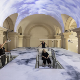 Ein Saal mit großen Säulen auf dessen Decke und Boden Wolken projiziert sind.