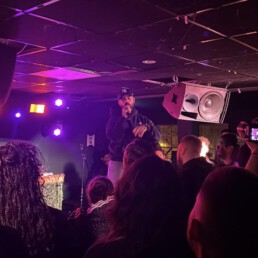 Der Rapper steht mit Mikrofon in der Hand auf einer kleinen Clubbühne in unmittelbarer Nähe zur Crowd.
