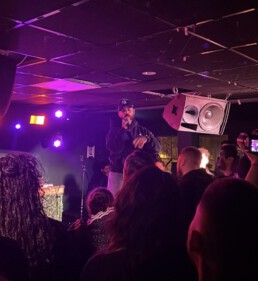 Der Rapper steht mit Mikrofon in der Hand auf einer kleinen Clubbühne in unmittelbarer Nähe zur Crowd.