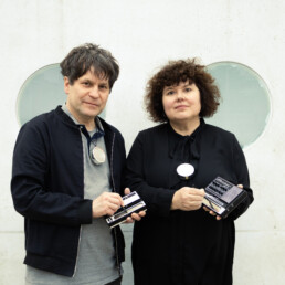 Silvana Battisti und Marc Herbert stehen mit elektronischen Instrumenten nebeneinander vor einer Wand