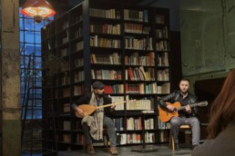 zwei Musiker auf einer B&uuml;hne vor einem gro&szlig;en B&uuml;cherregal